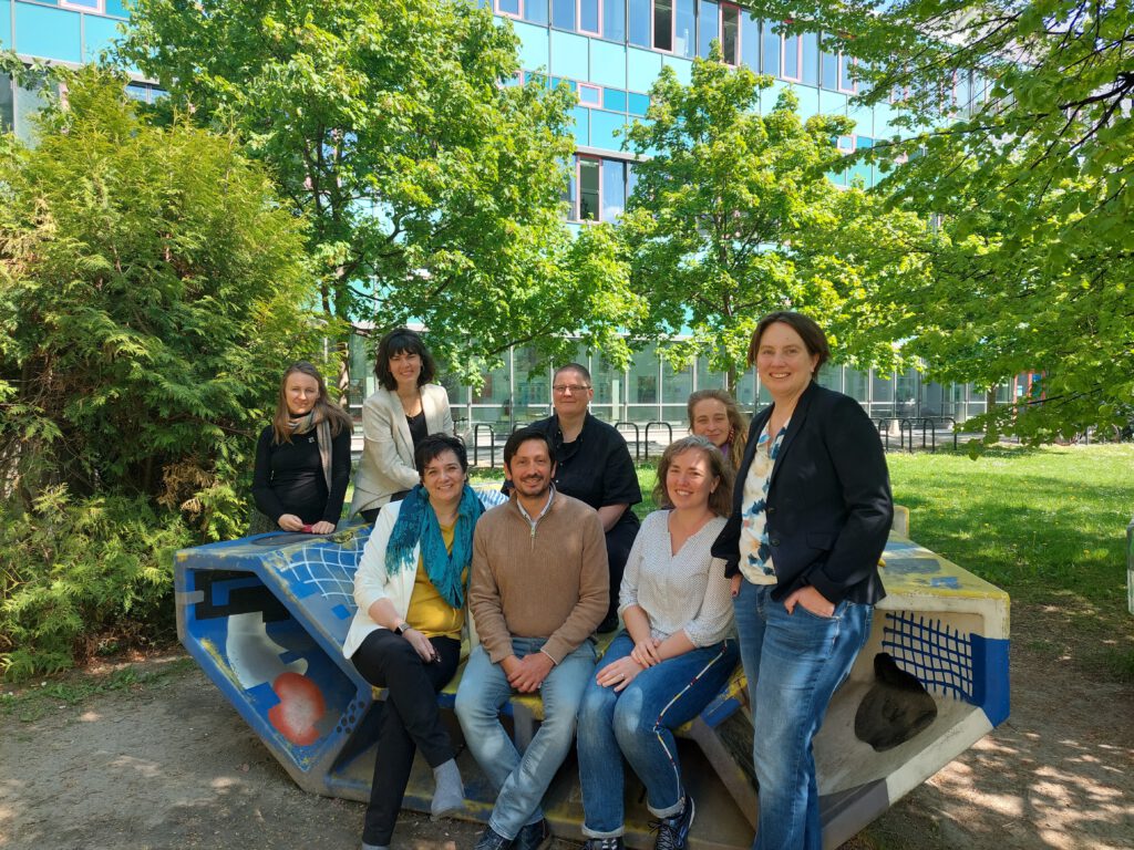 Group photo. Six women, a man, and a non-binary person sit on or stand around a colorfully painted concrete object. Trees & a Vienna University building in the background. 