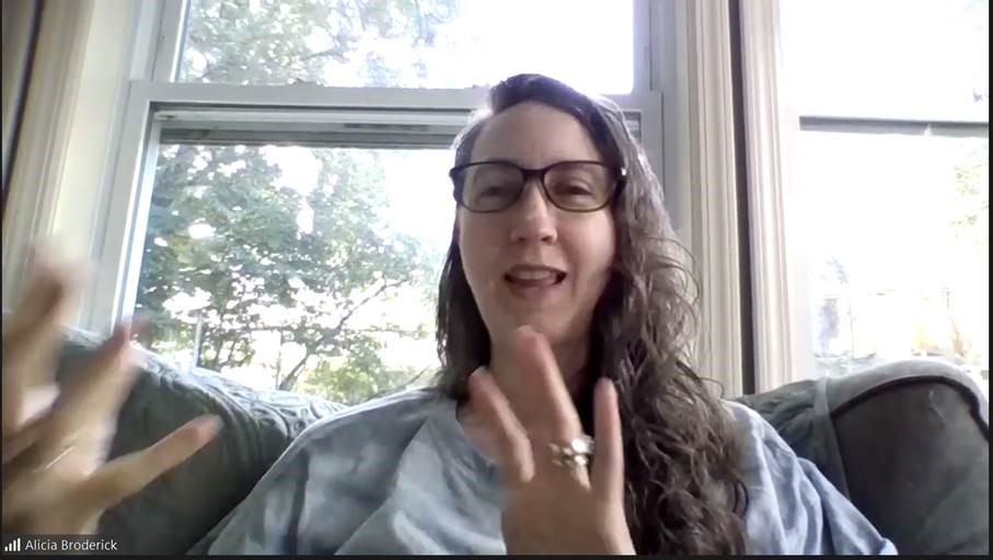 Alicia Broderick presenting via Zoom. You can see a woman with long, slightly curly hair, glasses and a light blue top. She is sitting on a sofa or armchair, looking at the camera, smiling while speaking and gesturing. In the background, you can see a tree through a large window.