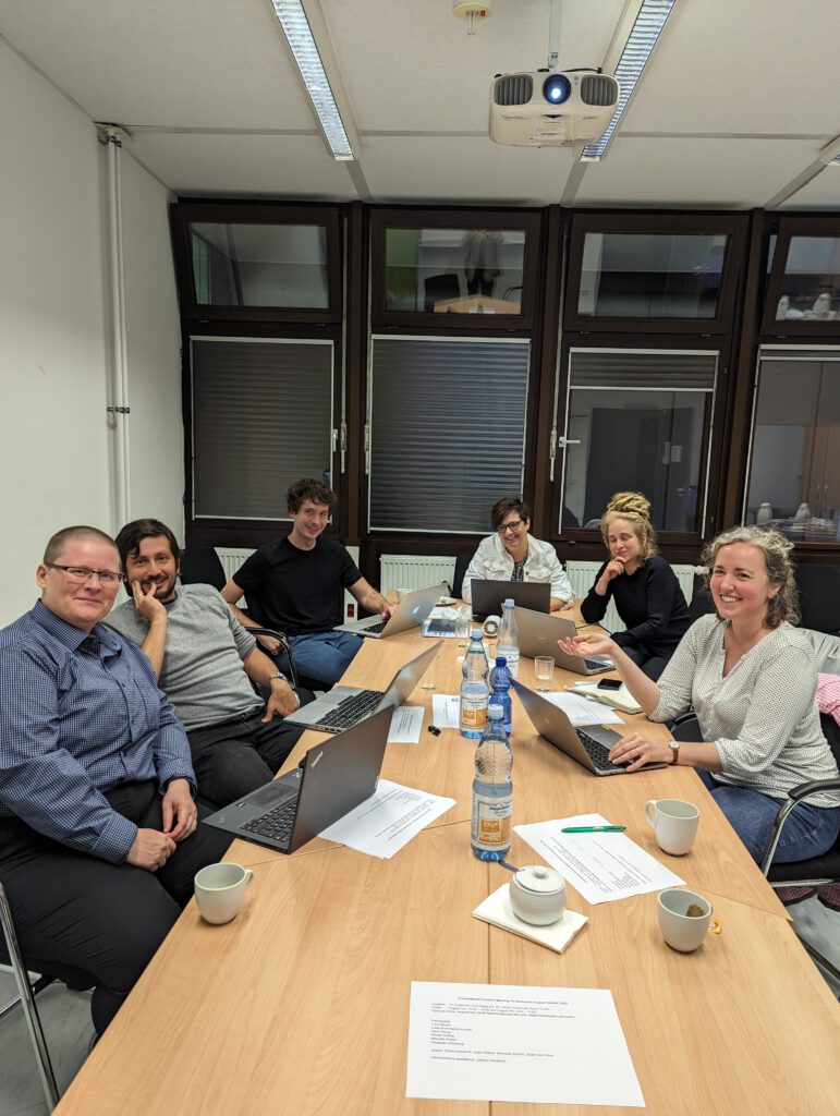 Three women, two men and a non-binary person sitting around a conference table, looking at the camera and smiling On the conference table, there are laptops, documents, coffee cups, etc. 