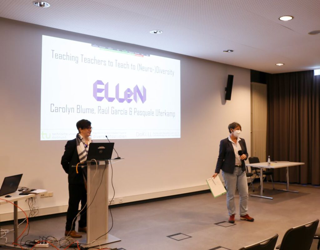 Two women in front of a powerpoint presentation. Caro Blume has short hair, wears glasses, black clothes and a grey scarf and is standing behind a lectern. Julia Hütner is on the right of the picture, with short hair, a mask, white shirt, black jacket and blue jeans. She is holding a microphone and a clipboard.