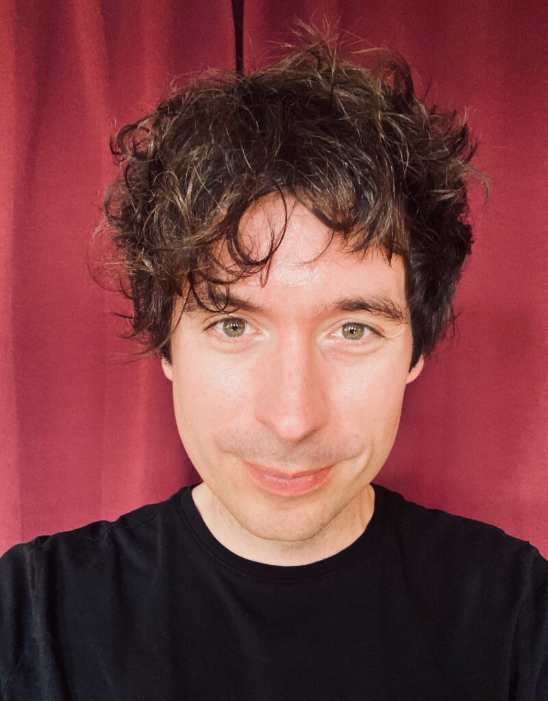 A portrait photo of a white person with fuzzy hair. He wears a black shirt.